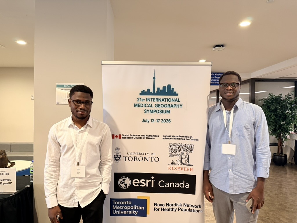 Two lab members beside the banner for the 21st International Medical Geography Symposium, Toronto, July 2026