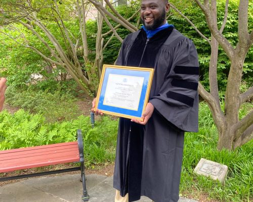 Dr. Meshack Achore in academic regalia holding his framed Lawrence A. Stessin Prize certificate